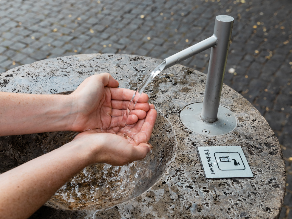 Auf dem Bild: Ein Trinkwasserbrunnen und zwei Hände, die das Wasser auffangen.